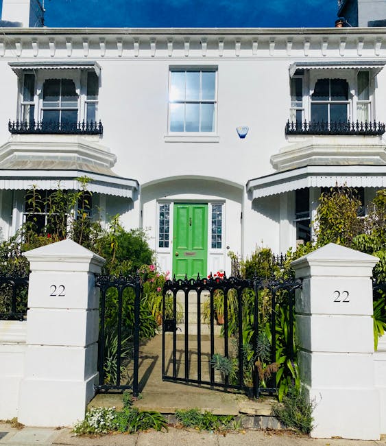 A white Victorian-style house with decorative black railings on the small balconies above the windows, featuring a vibrant green front door centered between two white pillars. Surrounding the entrance