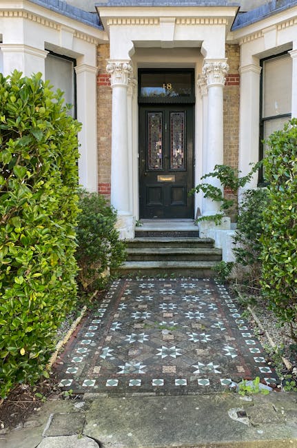 A white Victorian-style house with decorative black railings on the small balconies above the windows, featuring a vibrant green front door centered between two white pillars. Surrounding the entrance